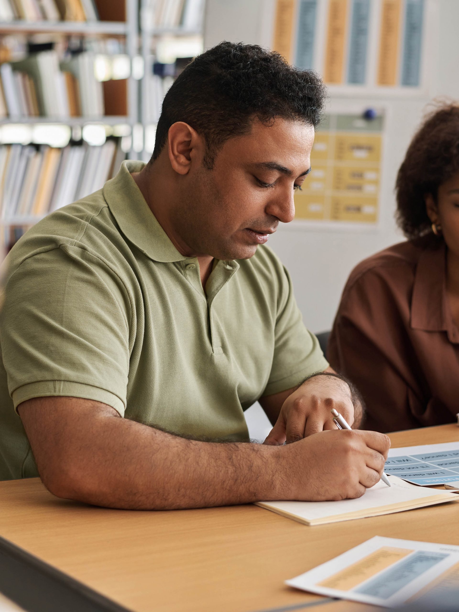 En medelålders man och yngre kvinna med olika bakgrund sitter vid ett bord och studerar i ett bibliotek på en kurs för att lära sig svenska.