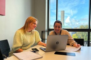 Två kvinnor som sitter framför en dator, med anteckningar och ett CV i handen i ett rum med utsikt över Stockholm – från CV och jobbcoaching för asylsökande hos Sensus.