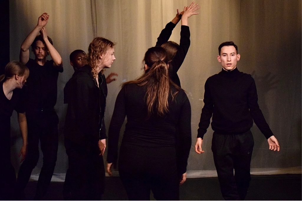 Flera svartklädda, unga vuxna/ungdomar i olika rörelser och i en dramatiskt ljussatt bild mitt under en teaterövning på Teater Aros på Midsommargården.
