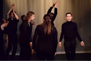 Flera svartklädda, unga vuxna/ungdomar i olika rörelser och i en dramatiskt ljussatt bild mitt under en teaterövning på Teater Aros på Midsommargården.