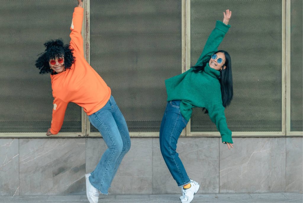 Två unga tjejer med färgglada tröjor mitt i en koordinerad dansrörelse. Bild för StudioUngs kostnadsfria aktiviteter / TIA-aktiviteter och möjlighet att dansa street dance gratis för alla mellan 16-25 år i Stockholm.