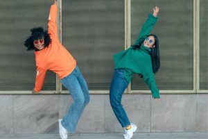 Två unga tjejer med färgglada tröjor mitt i en koordinerad dansrörelse. Bild för StudioUngs kostnadsfria aktiviteter / TIA-aktiviteter och möjlighet att dansa street dance gratis för alla mellan 16-25 år i Stockholm.