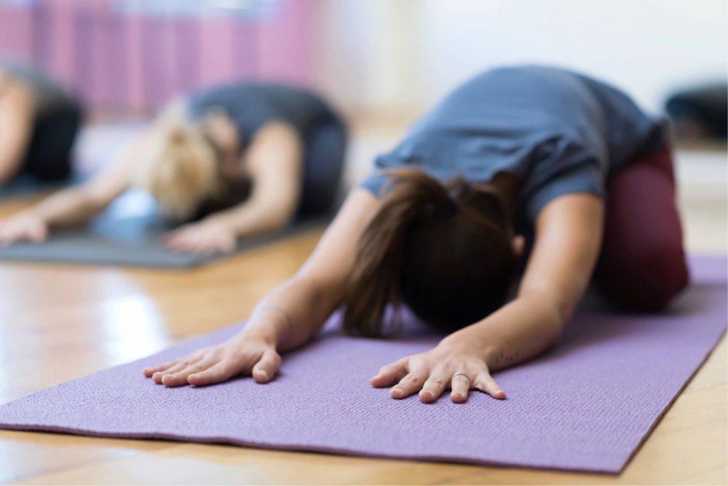 En kvinna i förgrunden, och flera andra personer i oskärpa i bakgrunden, där alla ligger i en yogaposition/stretchövning på yogamattor i en ljus sal.