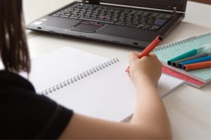 Närbild av hand som gör anteckningar framför datorn. Bild för Studiefrämjandets TIA-aktivitet fortsättningskurs i svenska på distans för asylsökande och vissa massflyktingar.