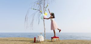 En kvinna står på en pall mot ett träd vid havet i bakgrunden. I en vig rörelse sträcker hon sig upp mot grenarna i trädkronan, som hon pyntar med färggrana ballonger.