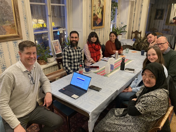 Åtta personer i olika åldrar och med olika bakgrund sitter runt ett bord och tittar in och ler mot kameran under en språkundervisning "Lär dig svenska för vardagen – Roligt och enkelt i Tensta!"