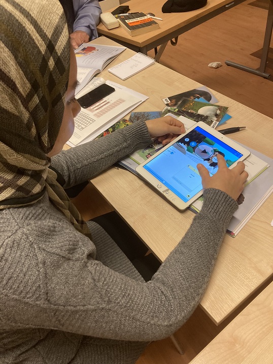 En kvinna sitter och gör övningar i svenska språket på en i pad i klassrumsmiljö, med mycket studiematerial utstpritt över borden runt henne.