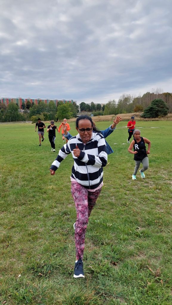 Olika personer i olika åldrar och från olika bakgrunder springer lekfullt och intensivt över en stor gräsmatta med höghus i bakgrunden. Bild från Inlöparna Ringeby RUN – långpass och söndagspromenad i Akalla.