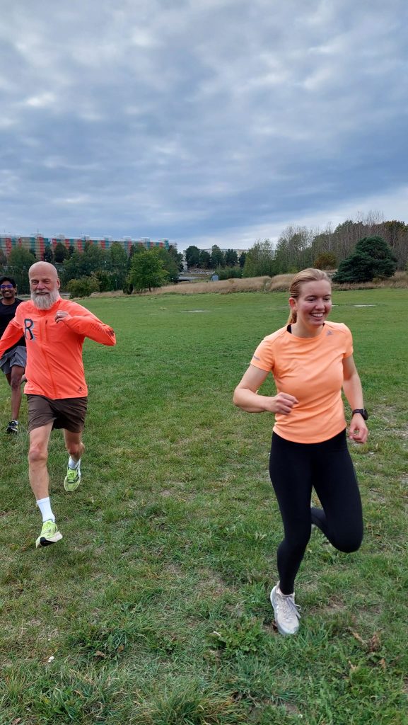 Tre personer i olika åldrar och från olika bakgrunder springer lekfullt och intensivt över en stor gräsmatta med höghus i bakgrunden. Bild från Inlöparna Rinkeby RUN – långpass och söndagspromenad i Akalla.