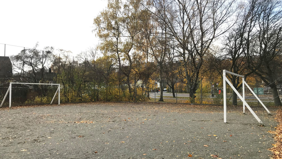 En tom fotbollsplan i grus med höstlöv och träd i höstfärger i bakgrunden.