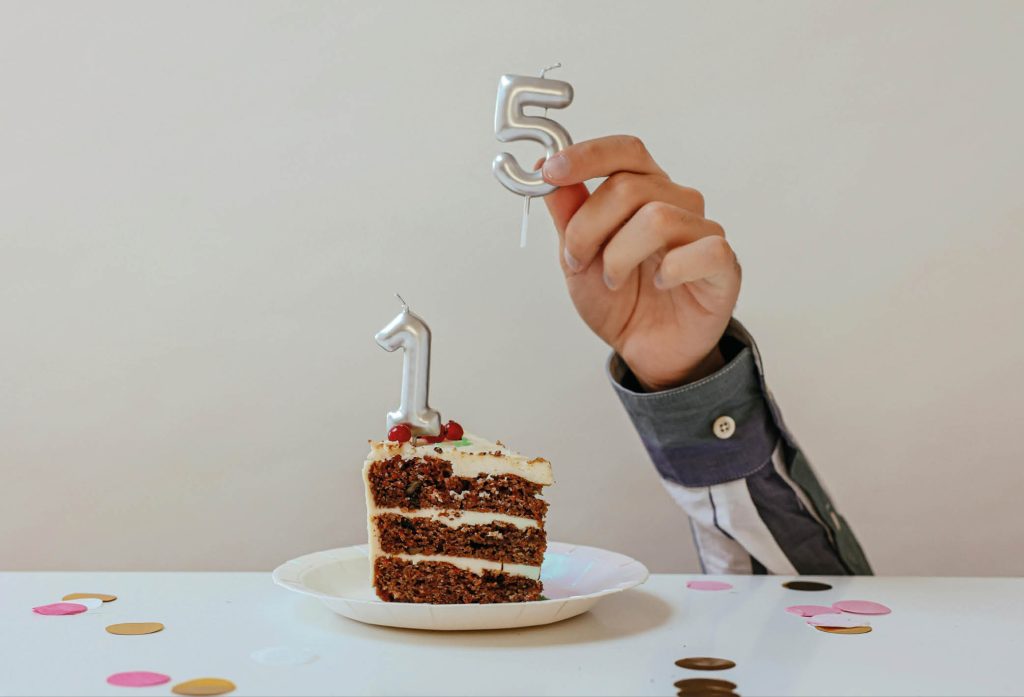 En tårtbit på ett fat, upplagt på ett festligt dekorerat bord med konfetti. I tårtbiten sitter ett ljus med siffran "1", och intill den bakom bordet sträcker sig en liten hand upp som håller i ett ljus strax bredvid ettan, fast med siffran "5". Tillsammans ser man att det blir siffran 15.